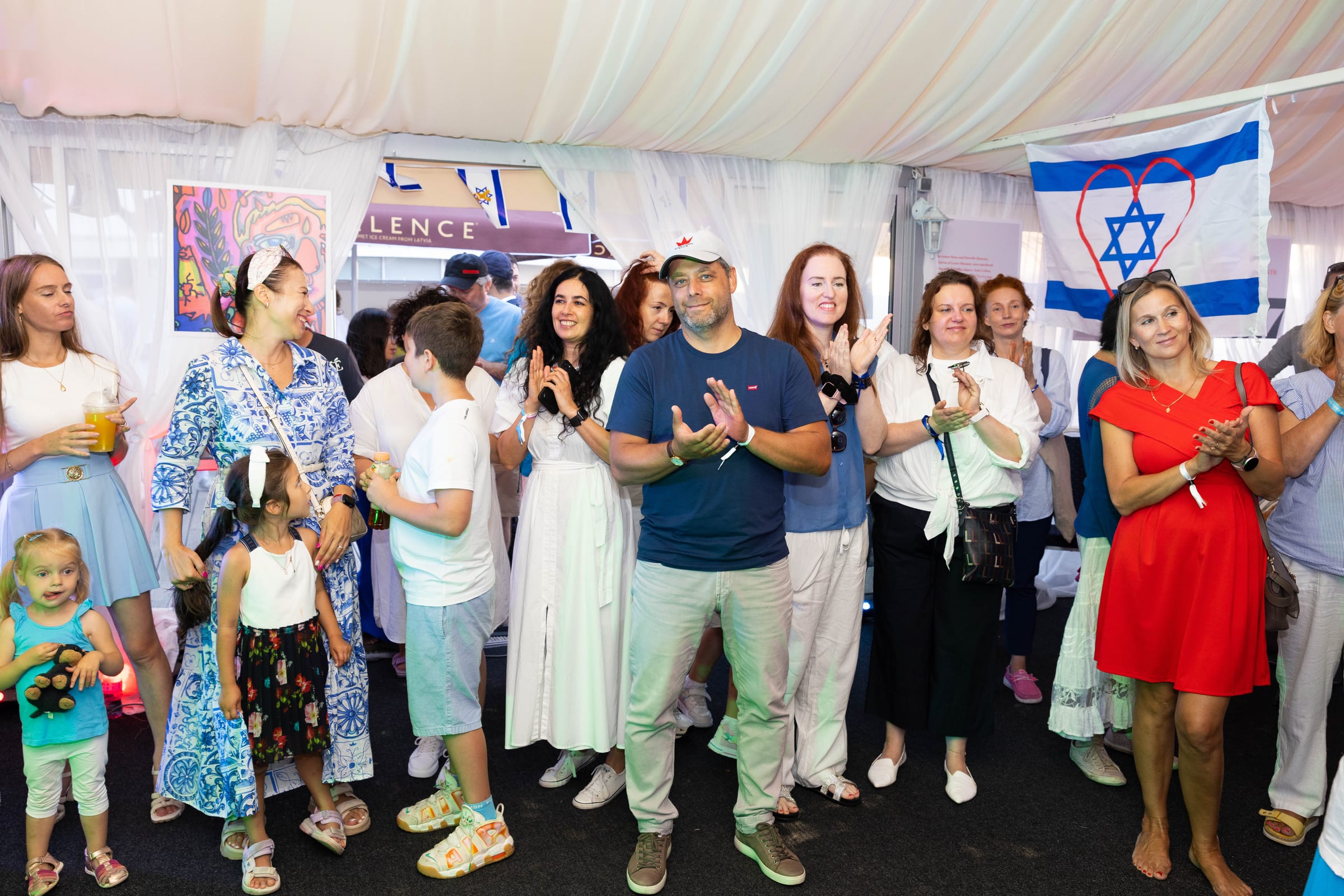 Community applauding inside a tribute tent with an Israeli flag